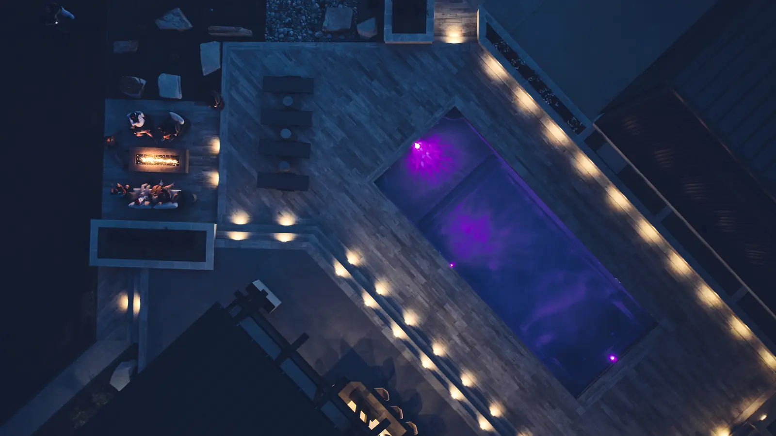 Night aerial of an LED-lit pool glowing beside a burning fire pit lounge at the Johnson Estate