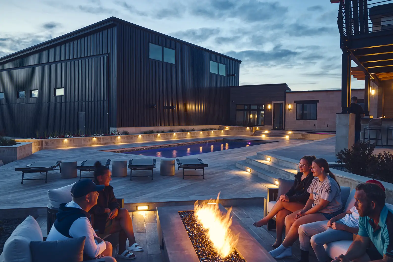 Family gathered around the fire pit beside the lit pool at night at the Johnson Estate